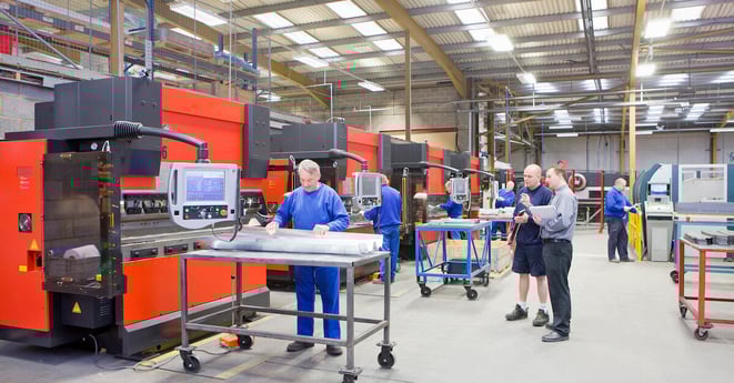 A wide shot of the workers working on the machines while a managers watches them in a factory.