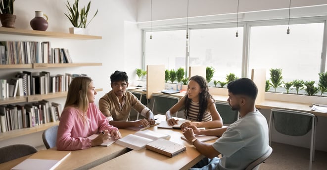 Four students sitting at table in university classroom discussing topics related to studies or joint project completion, researching, preparing for exams or admission. Team-work, knowledge exchange