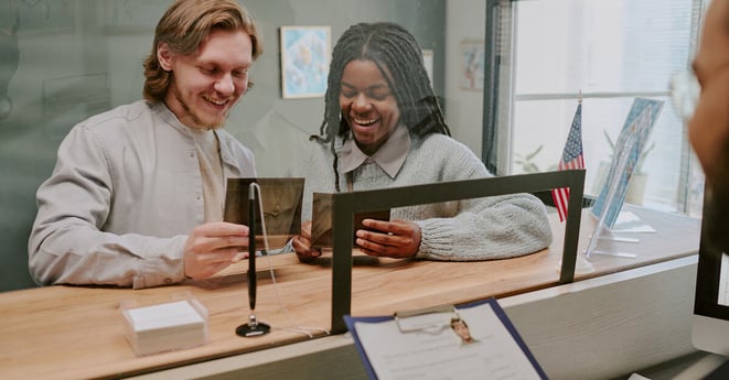 Smiling couple sitting at counter of public office, filling out forms and holding ID cards while engaging with official sitting behind glass partition