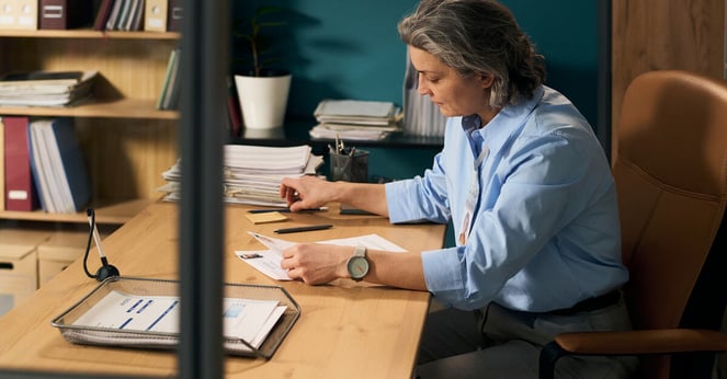 Middle aged Caucasian woman social worker sitting at desk reviewing paperwork in organized office environment, demonstrating focus and professionalism in social services setting