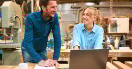 Male And Female Carpenters Working In Woodwork Workshop Using Laptop Together