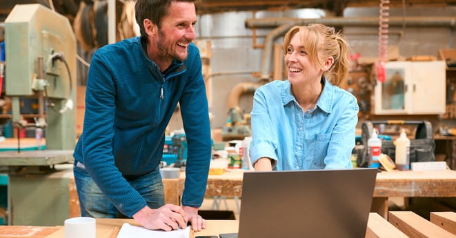 Male And Female Carpenters Working In Woodwork Workshop Using Laptop Together