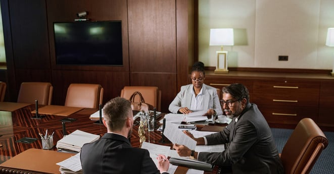 Group of intercultural coworking lawyers in formalwear sitting by large table in boardroom and working with paper documents
