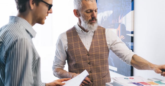 Senior Caucasian businessman in checkered vest helps younger male colleague with document printing at office printer. They analyze charts and collaborate on preparing colorful printed materials