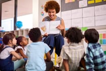 Female Teacher Reads To Multi-Cultural Elementary School Pupils Sitting On Floor In Class At School