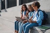 Side view of happy group of multiethnic school kids preteen boys and girls elementary middle pupils children sitting on the stairs and using digital devices laptop tablet smartphone outdoors