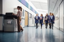 Group of teenage girls are walking down the school hall with books and laptops in their arms. They are talking and laughing as they walk and there is a female teacher using the printer.