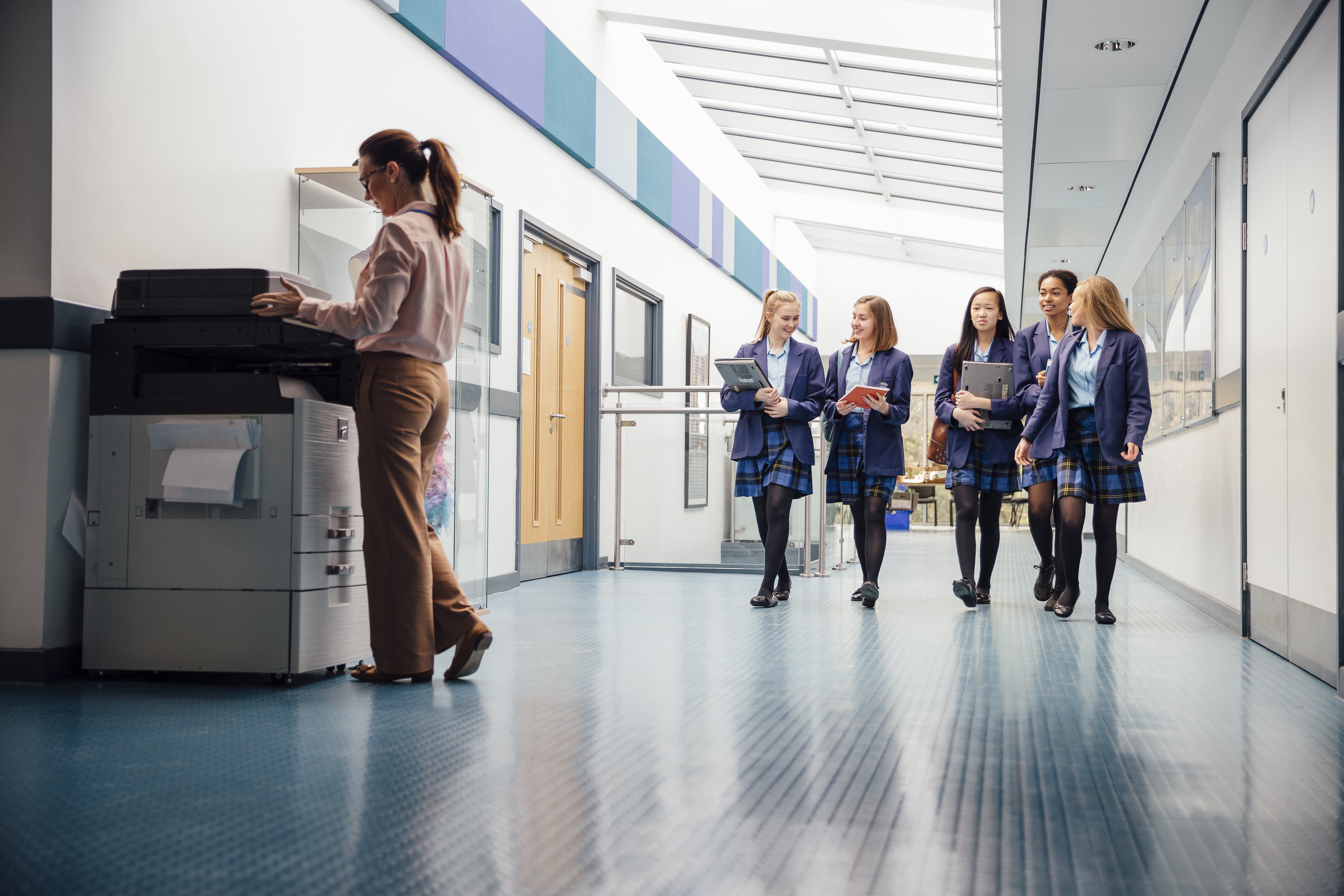 Group of teenage girls are walking down the school hall with books and laptops in their arms. They are talking and laughing as they walk and there is a female teacher using the printer.